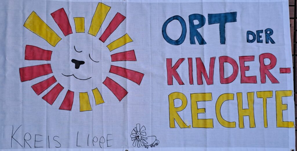 Zu sehen ist ein Banner vom Ort der Kinderrechte. Kinder aus dem Kreis Lippe haben das Löwen-Logo und den Schriftzug "Ort der Kinderrechte" für die Eröffnungsfeier gemalt.